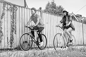 Feel Good Wealth Your Be Ethical with a man and woman each riding a bicycle in front of a wooden fence.